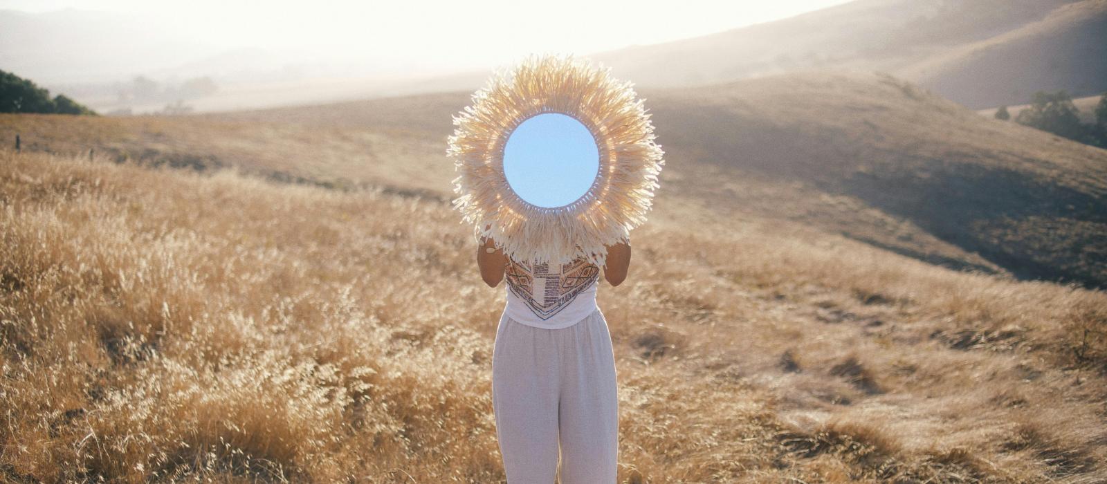 Kobieta na tle wzgórz trzyma lustro zakrywając twarz