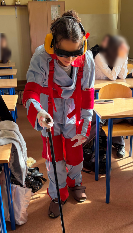 osoba w kombinezonie geriatrycznym spaceruje po klasie o lasce