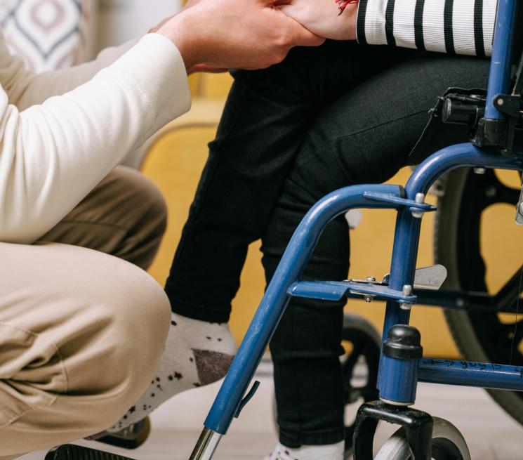 zdjęcie dwie osoby jedna siedządza na wózku inwalidzkim i kucający przy niej mężczyzna trzymają się za ręce