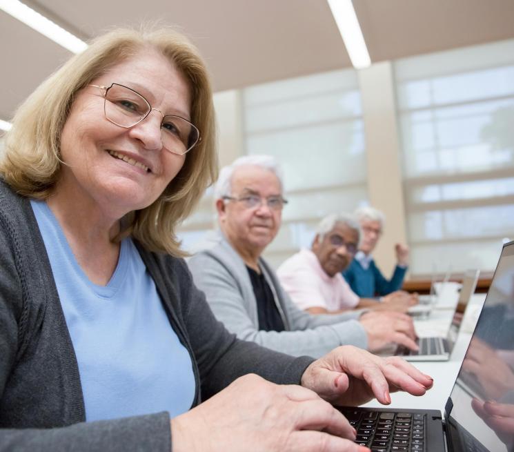 zdjęcie kilku seniorów siedzi przy biurkach i pracuje na laptopach uśmiechają się