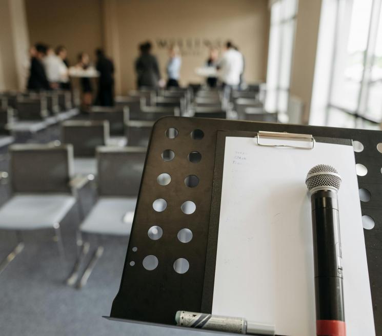 zdjęcie sala konferencyjna na pierwszym planie mównica z mikrofonem i białą kartką w tle rozmyci uczestnicy