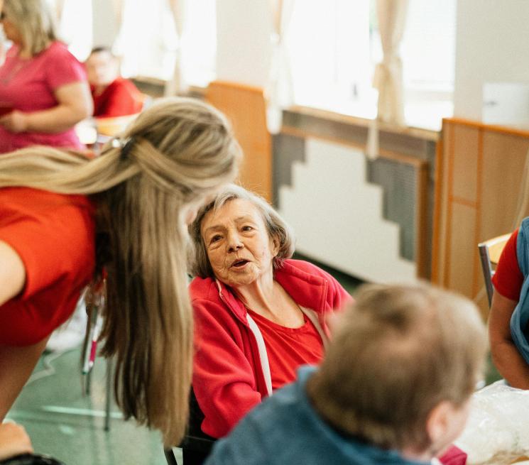 zdjęcie opiekunka schyla się i mówi coś do starszej kobiety siedzącej na wózku inwalidzkim