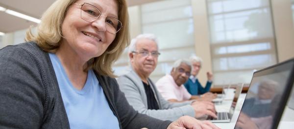 zdjęcie 4 uśmiechniętych seniorów w rzędzie siedzą przy stołach przy laptopach 