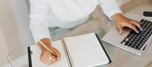 zdjęcie kobieta w białej koszuli siedzi przy biurku pracuje na laptopie i notuje w kalendarzu