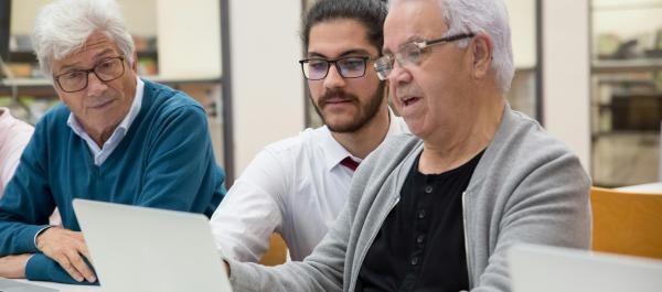 zdjęcie dwóch starszych mężczyzn pracuje przy laptopach pomiędzy nimi kuca młodszy meżczyzna i coś im tłumaczy