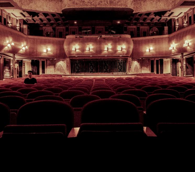 zdjęcie wnętrza teatru rzędy bordowych krzeseł na jednym z krzeseł po lewej stronie siedzi młody mężczyzna