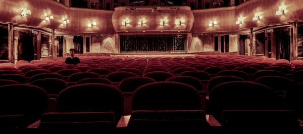 zdjęcie wnętrza teatru rzędy bordowych krzeseł na jednym z krzeseł po lewej stronie siedzi młody mężczyzna