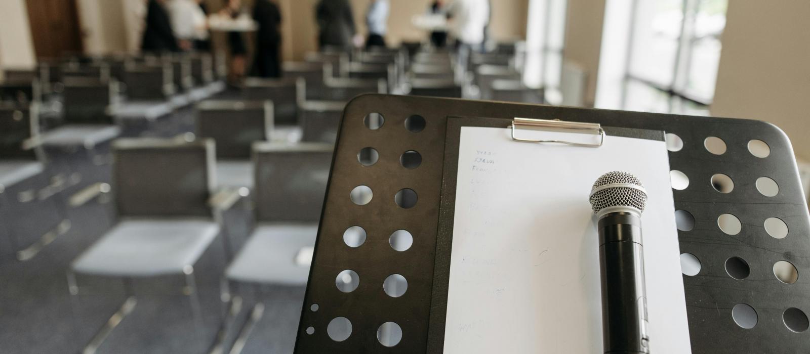 zdjęcie sala konferencyjna na pierwszym planie mównica na niej leży biała kartka oraz mikrofon w tle kilka osób 